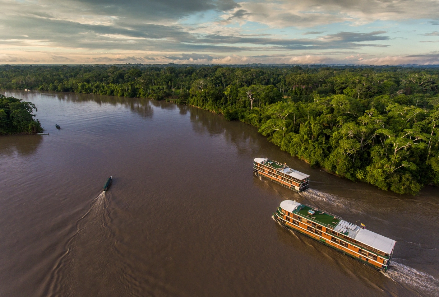 Aerial view of two flotels cruising the wide, brown Napo River in Ecuador, surrounded by lush green rainforest. Sunset at the horizon.