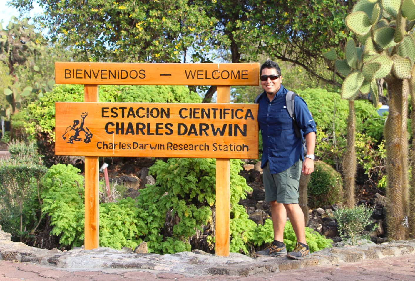 Adult male traveler posing with the sign at the entrance of Charles Darwin Station in Galapagos. The wooden sign reads Welcome, Charles Darwin Research Station in English and Spanish. Lush green vegetation surrounds the scene.